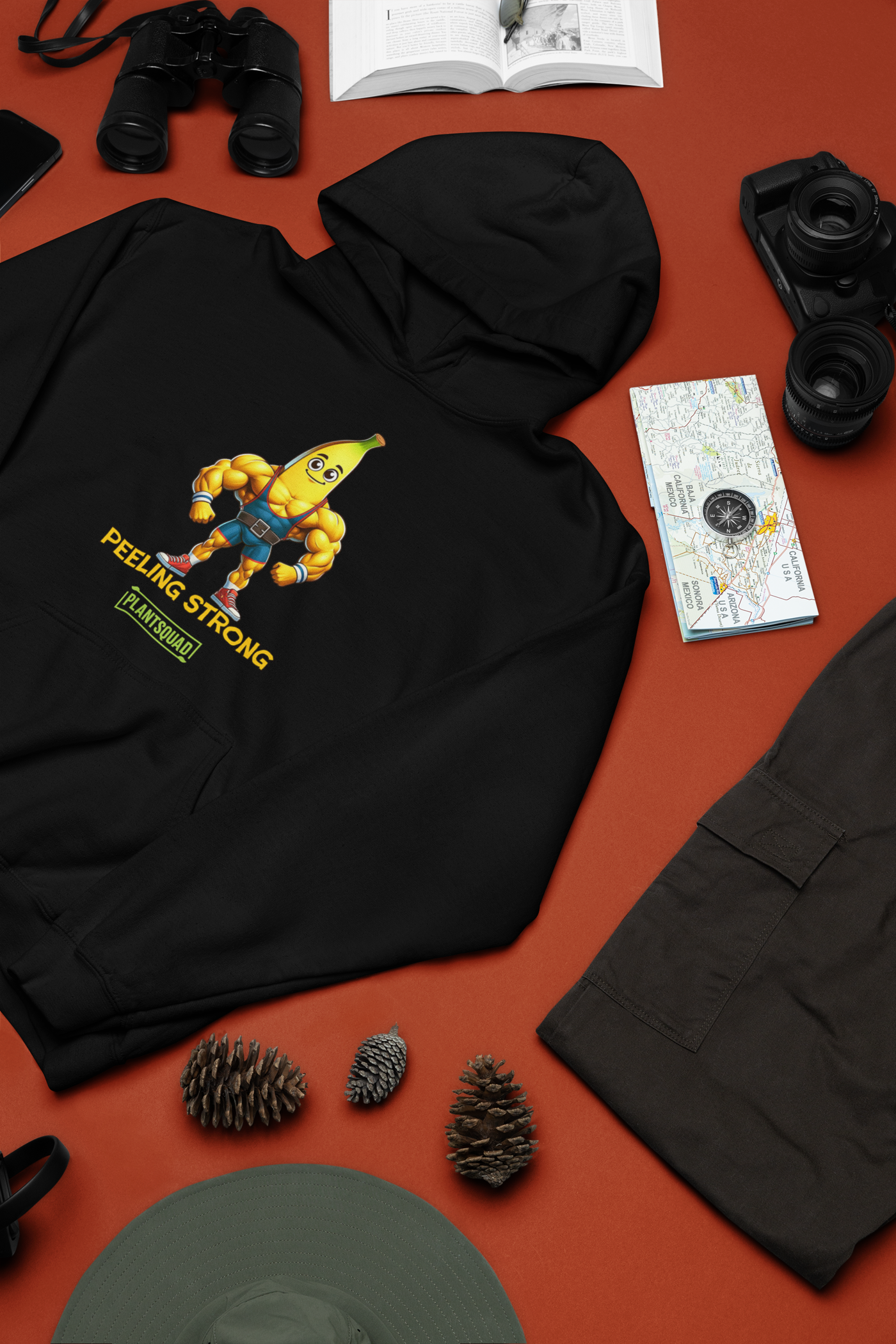 A Plantsquad Banana "Peeling Strong" - Unisex Hoodie features a cartoon banana with muscular arms and legs, wearing shorts and a weightlifting belt. The text reads "PEELING STRONG" above "PLANTSQUAD," perfect for those embracing a vegan lifestyle.