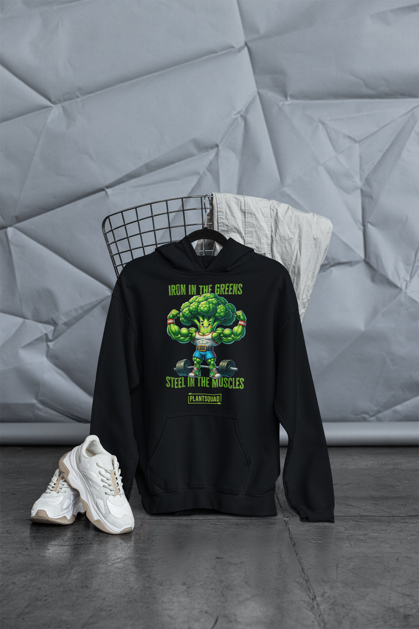 A Plantsquad Broccoli "Iron In The Greens Steel In The Muscles" - Unisex Hoodie featuring a muscular cartoon broccoli character lifting dumbbells, with the text "Iron in the greens, steel in the muscles" above and "Plant Squad" below. Embrace your vegan lifestyle and love of weight lifting with this fun design. The character sports a determined expression, white tank top, and yellow shorts.