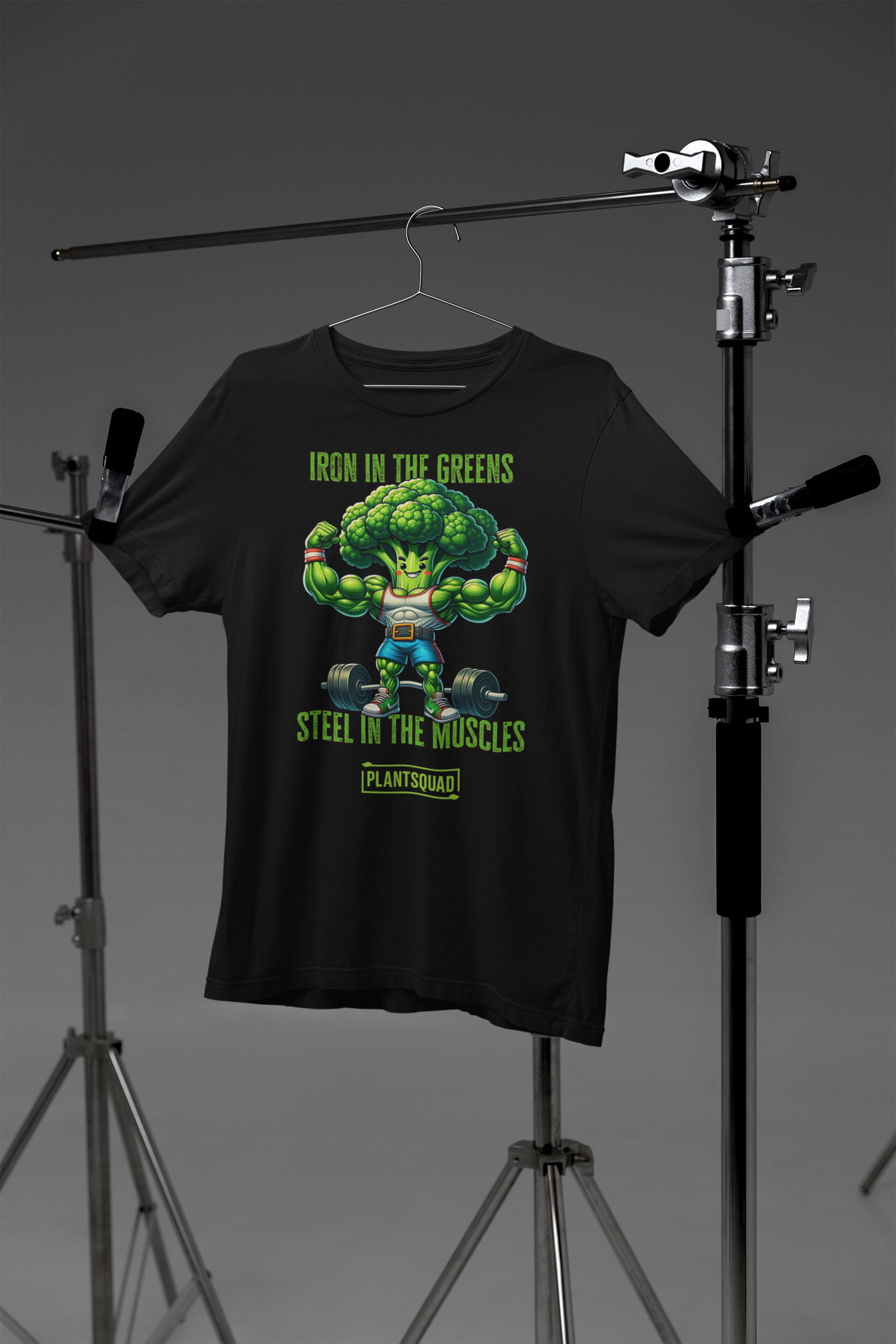 A black Plantsquad Broccoli "Iron In The Greens Steel In The Muscles" - Unisex T-Shirt features an illustrated muscular broccoli character lifting weights. Above the character, the text reads, "Iron in the greens." Below, it says, "Steel in the muscles." At the bottom, there's a bold "PLANTSQUAD" logo.