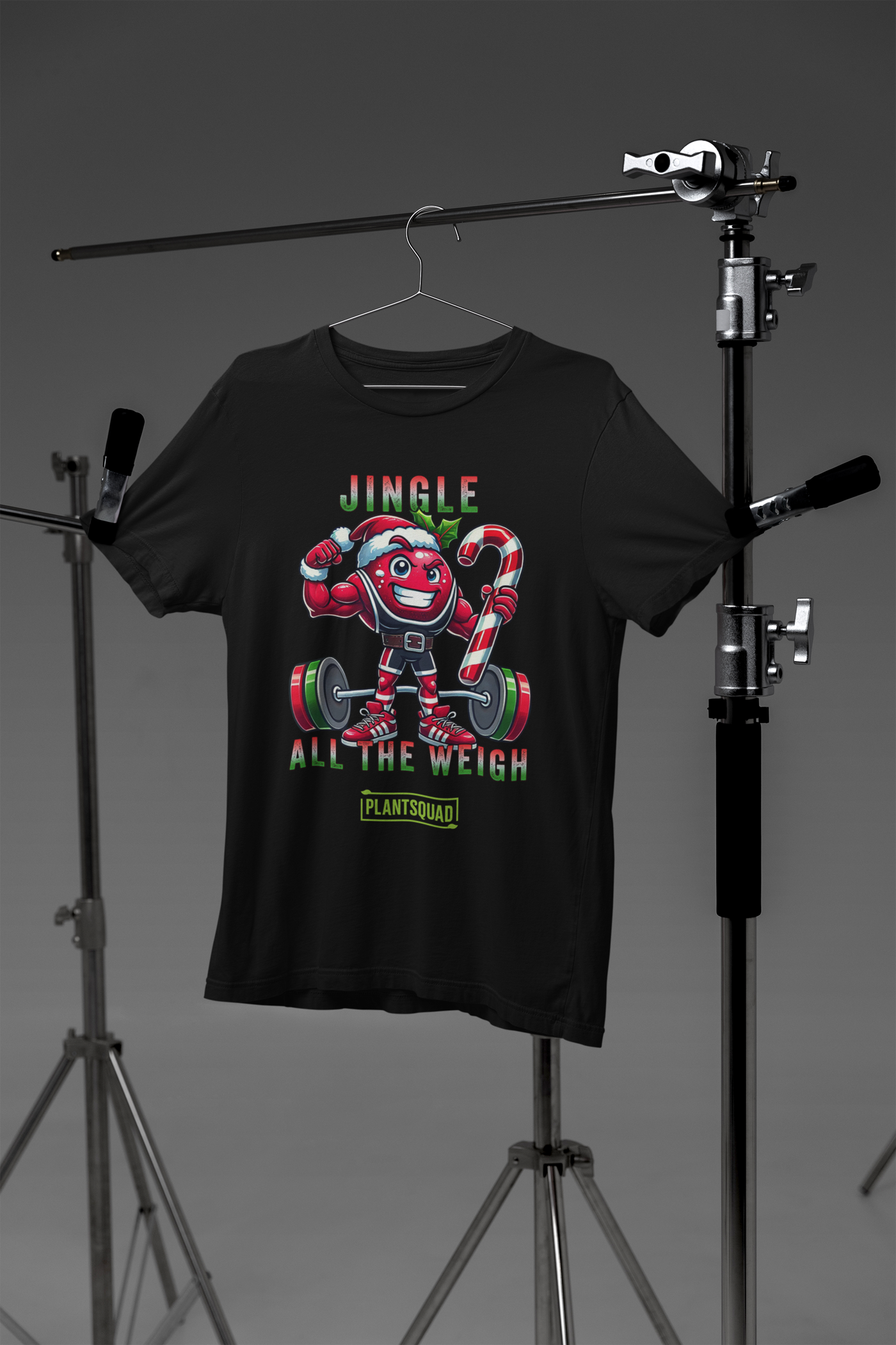 A picture of a t-shirt featuring a strong looking cranberry cartoon character flexing its arms holding a candy cane. A small green logo "PLANTSQUAD" appears above the character, "JINGLE ALL THE WEIGH" below. Perfect for those embracing a plant-based or vegan lifestyle that are into fitness