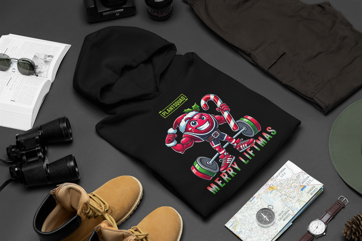 A folded t-shirt on a table with a muscled cranberry cartoon character flexing its arms holding a candy cane. A small green logo "PLANTSQUAD" appears above the character, "MERRY LIFTMAS" below. Perfect for those embracing a plant-based or vegan lifestyle that are into fitness