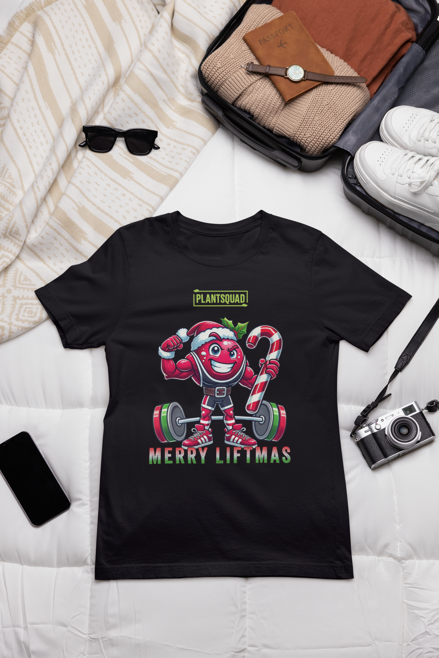 A t-shirt laying flat on a table with a muscled cranberry cartoon character flexing its arms holding a candy cane. A small green logo "PLANTSQUAD" appears above the character, "MERRY LIFTMAS" below. Perfect for those embracing a plant-based or vegan lifestyle that are into fitness
