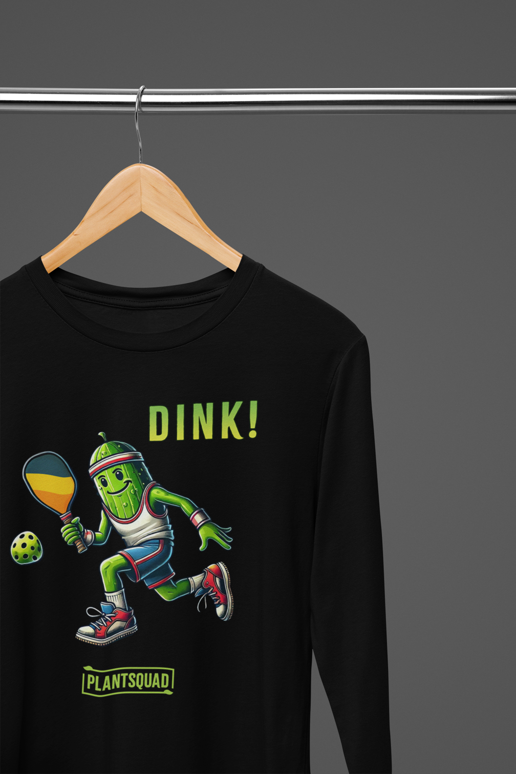 A hanging Long Sleeve T-shirt featuring a pickle playing pickleball. There is a caption listed as "Dink!", with a small green logo "PLANTSQUAD" underneath. Perfect for those who love pickleball.