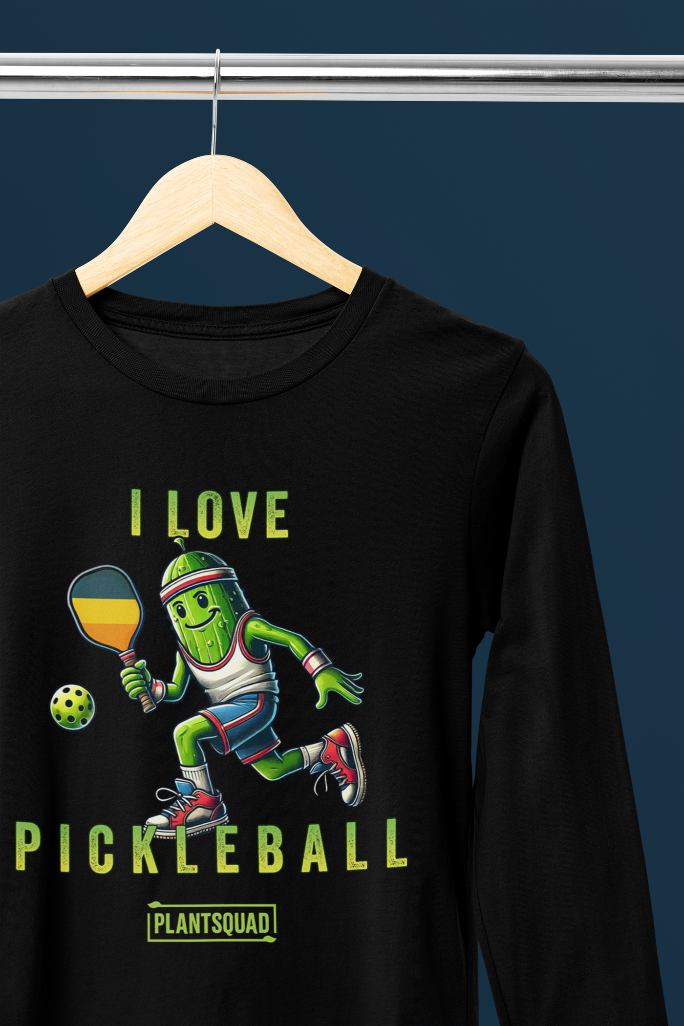 A hanging Long Sleeve T-shirt featuring a pickle playing pickleball. There is a caption listed as "I Love Pickleball", with a small green logo "PLANTSQUAD" underneath. Perfect for those who love pickleball.