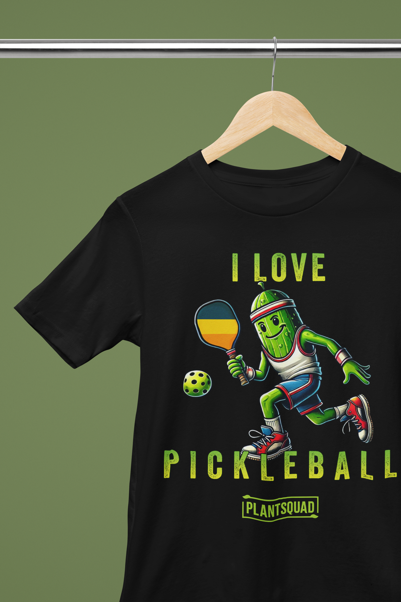 A hanging t-shirt featuring a pickle playing pickleball. There is a caption listed as "I Love Pickleball", with a small green logo "PLANTSQUAD" underneath. Perfect for those who love pickleball.