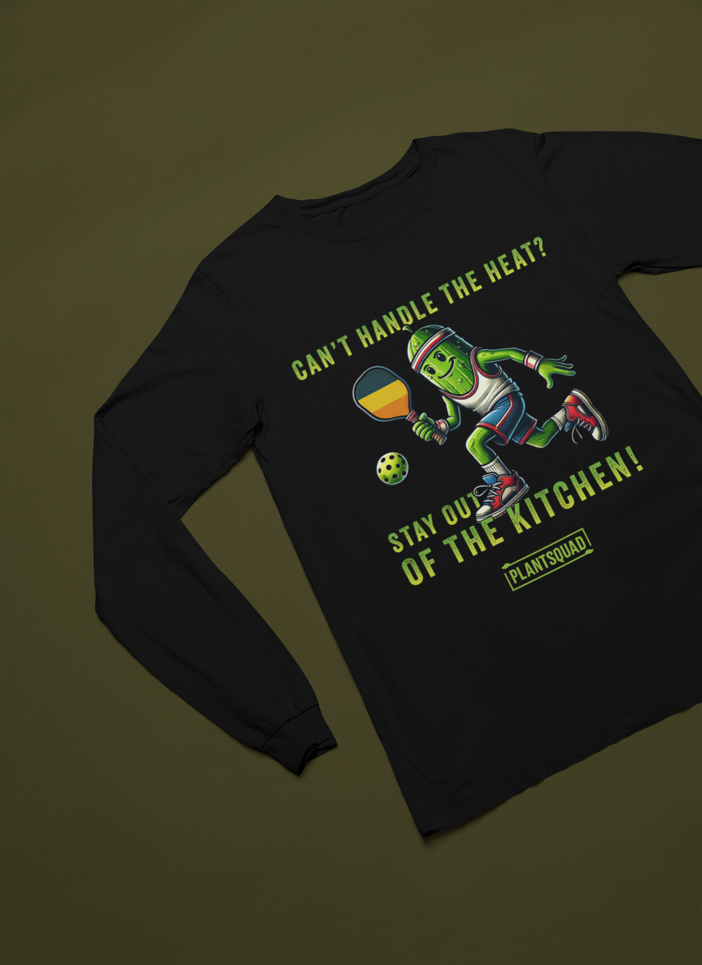 A picture of a laid out Long Sleeve T-Shirt featuring a pickle playing pickleball. There is a caption listed as "Can't Handle The Heat? Stay Out Of The Kitchen!", with a small green logo "PLANTSQUAD" underneath. Perfect for those who love pickleball.