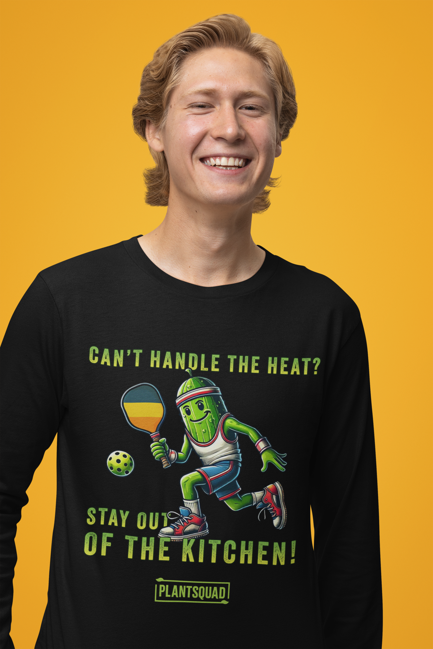 A model wearing a Long Sleeve T-Shirt featuring a pickle playing pickleball. There is a caption listed as "Can't Handle The Heat? Stay Out Of The Kitchen!", with a small green logo "PLANTSQUAD" underneath. Perfect for those who love pickleball.