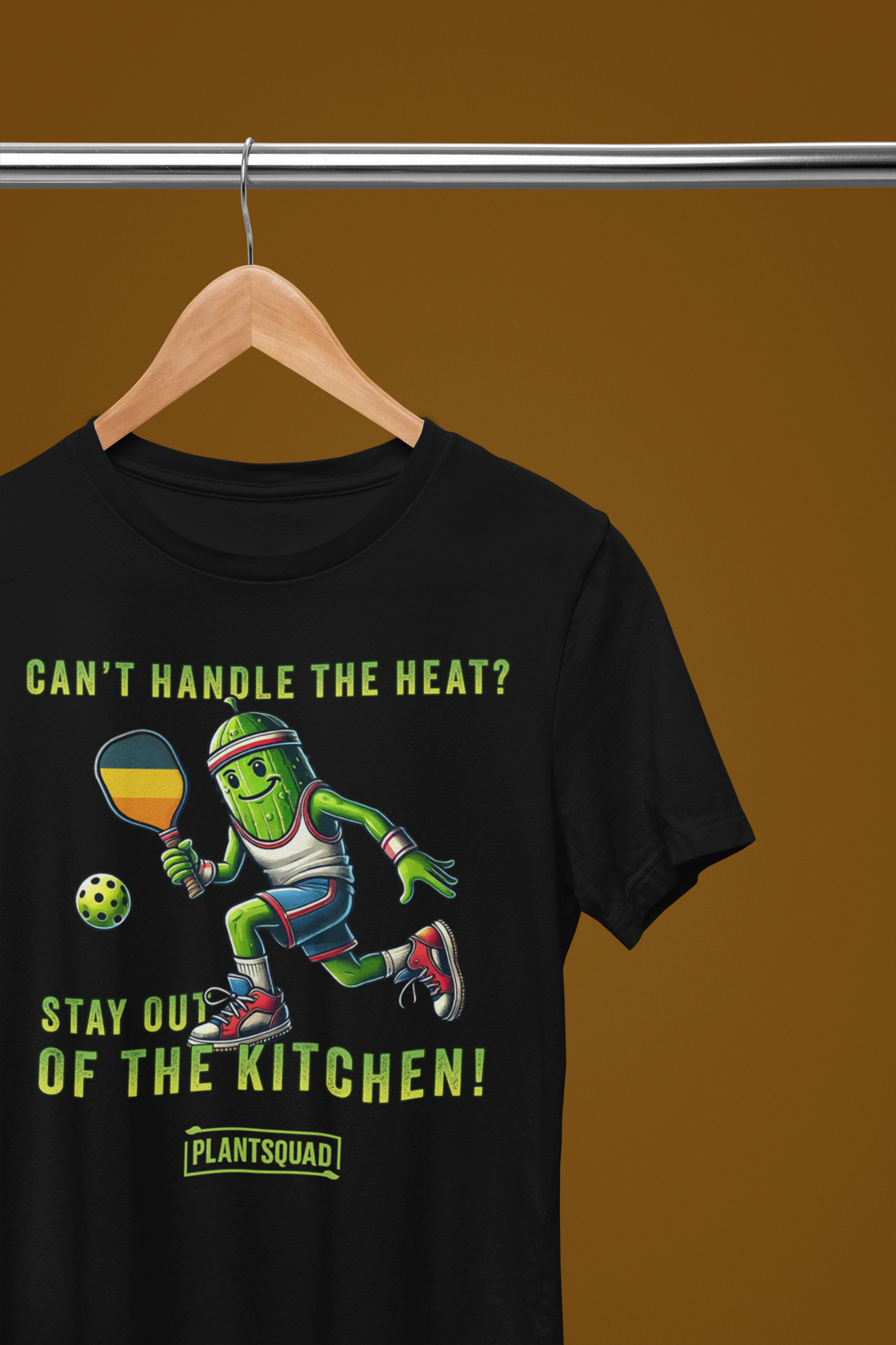A hanging t-shirt featuring a pickle playing pickleball. There is a caption listed as "Can't Handle The Heat? Stay Out Of The Kitchen!", with a small green logo "PLANTSQUAD" underneath. Perfect for those who love pickleball.