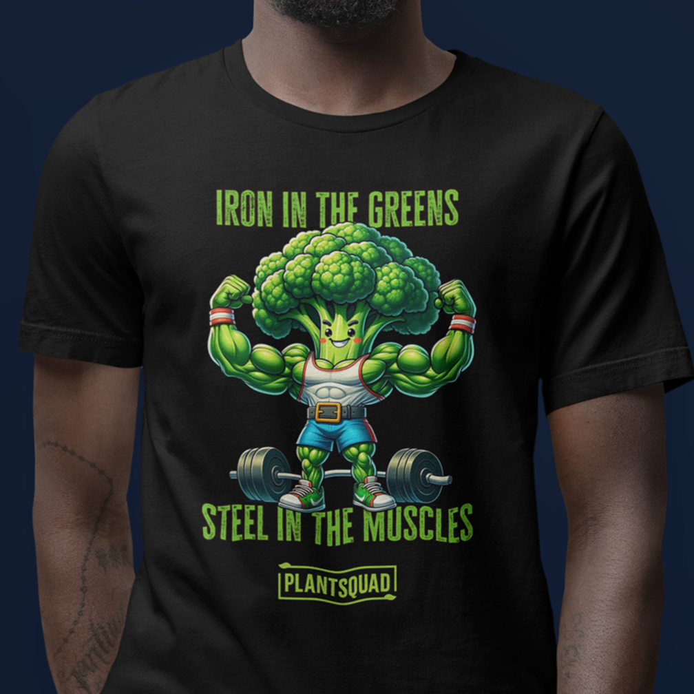 A person wears a black t-shirt featuring a cartoon broccoli character flexing muscles while lifting a barbell. The text reads "Iron in the Greens, Steel in the Muscles" above the character, with "PlantSquad" below it. The background is solid dark blue.