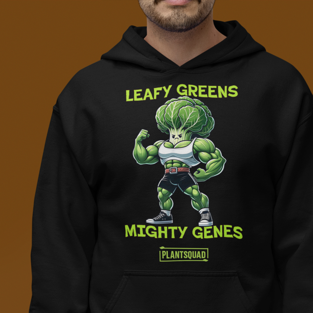 A person wearing a black hoodie featuring a cartoon muscular broccoli character flexing its biceps. The text above the character reads "LEAFY GREENS," and below it says "MIGHTY GENES." The bottom text says "PLANTSQUAD." The background is solid brown.