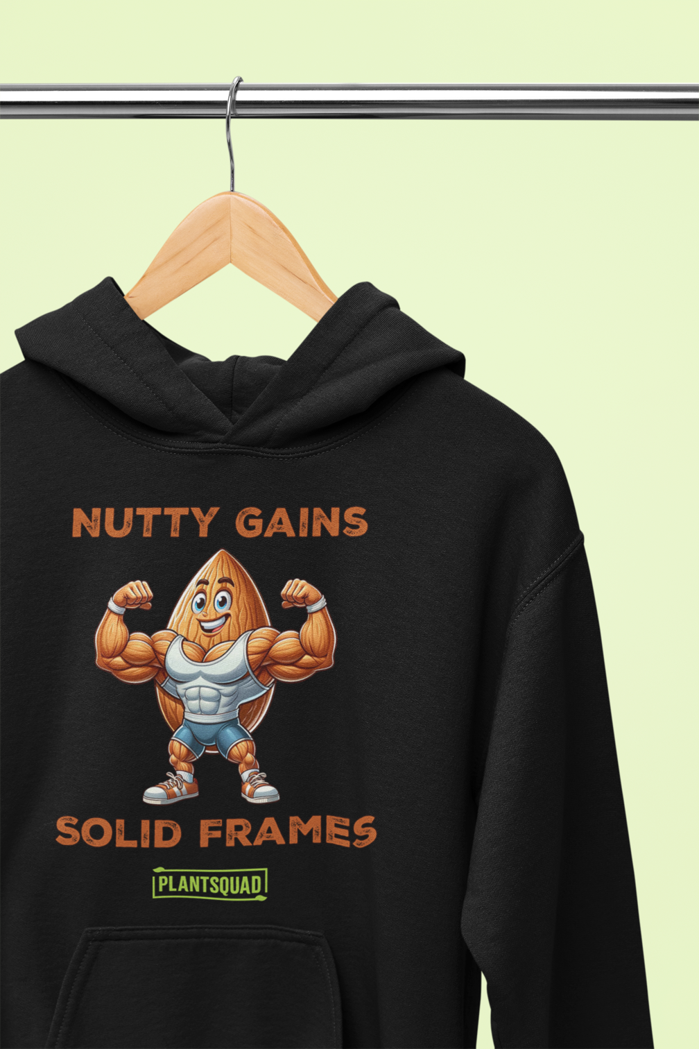 A black Plantsquad Almond "Nutty Gains Solid Frames" - Unisex Hoodie displays a muscular peanut character flexing its biceps with the text "Nutty Gains" above and "Solid Frames" below. The logo "Plant Squad" is at the bottom, perfect for gym enthusiasts. The design is bold, humorous, and fitness-themed.