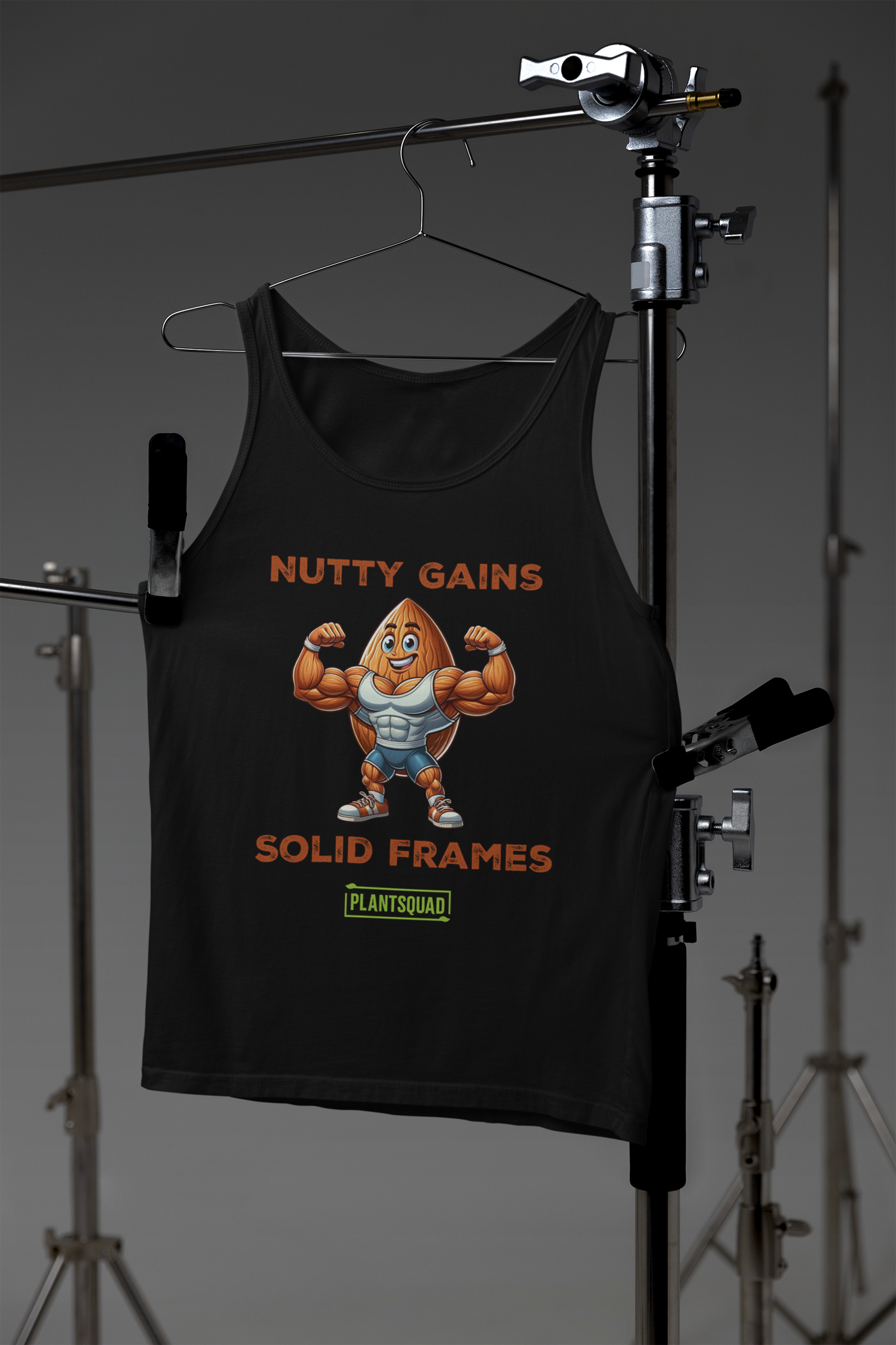 This Plantsquad Almond "Nutty Gains Solid Frames" - Unisex Tank Top features a buff, anthropomorphic almond flexing its muscles. Above it, in bold orange text, reads "NUTTY GAINS." Below, it says "SOLID FRAMES." At the bottom is a green rectangular logo with the text "PLANTSQUAD," perfect for showcasing your vegan lifestyle.