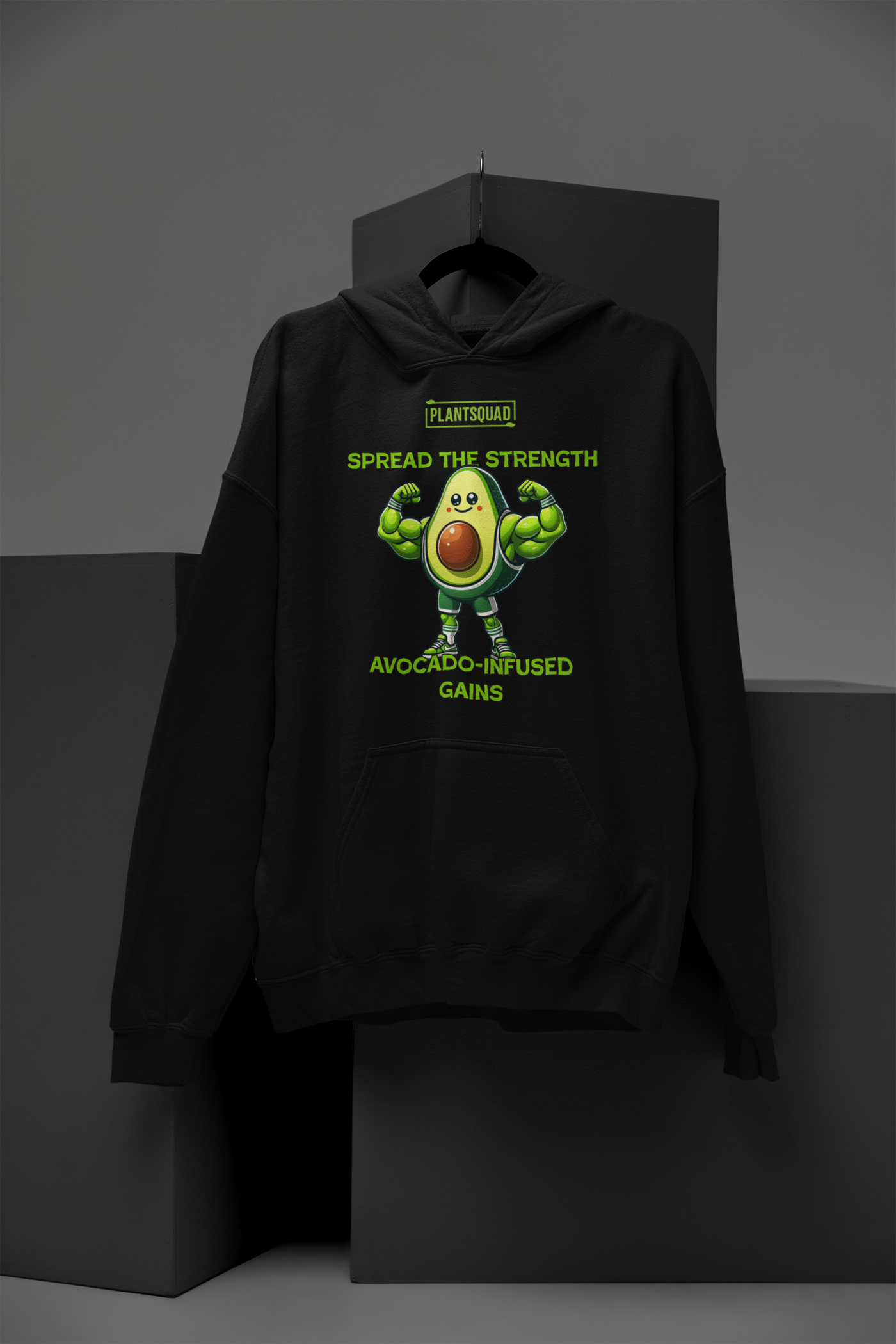 The **Plantsquad Avocado "Spread The Strength Avocado-Infused Gains" - Unisex Hoodie** features a smiling, muscular avocado character with flexed arms. Above it, the text reads "PLANTSQUAD." Below, in bright green letters: "SPREAD THE STRENGTH AVOCADO-INFUSED GAINS." Ideal for anyone embracing a vegan lifestyle and loving an avocado design.