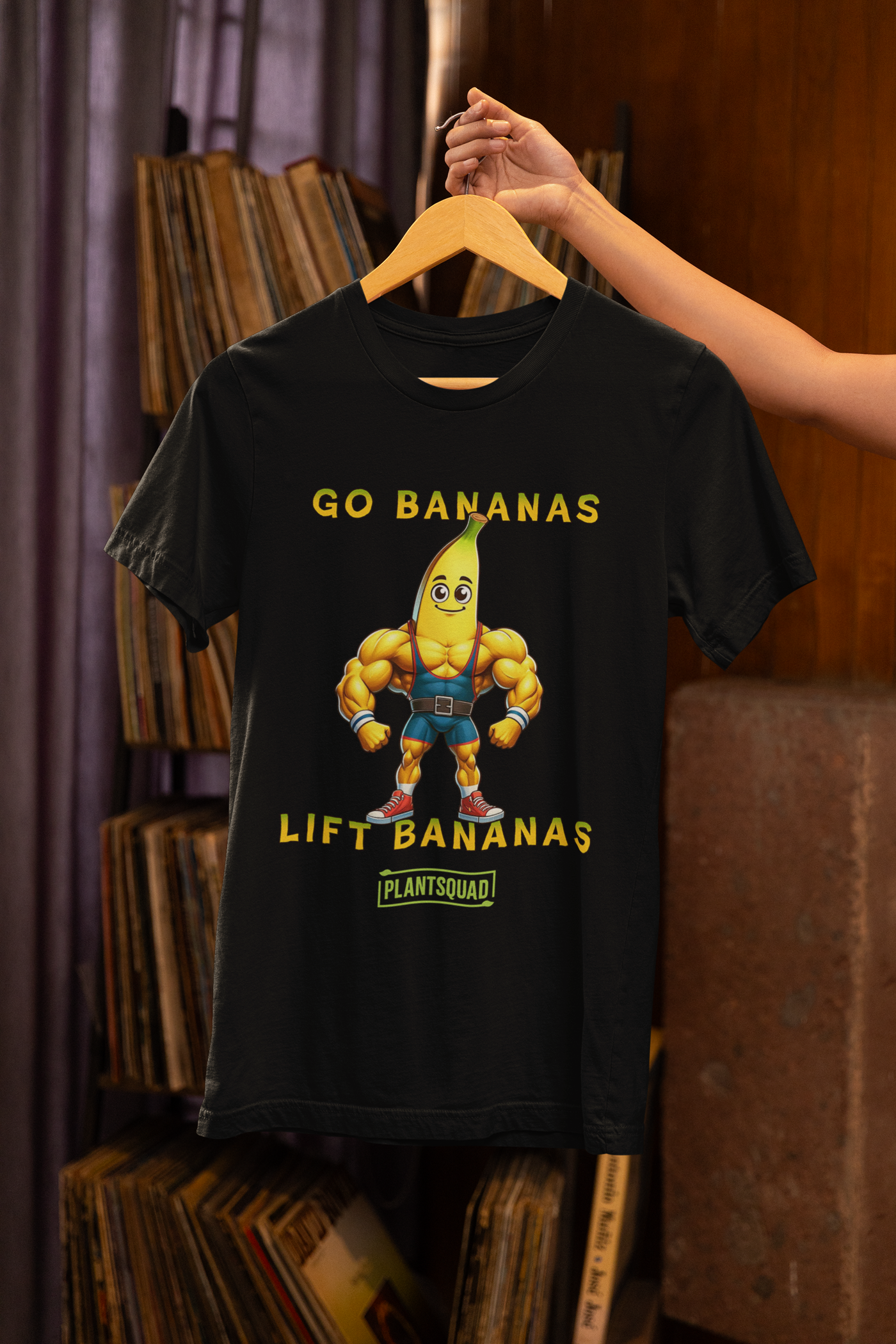 A Plantsquad Banana "Go Bananas Lift Bananas" - Unisex T-Shirt features a cartoon muscular banana wearing gym shorts, and boots. Above the banana, it says "GO BANANAS" and below it says "LIFT BANANAS". There is also a small green "plantsquad" logo at the bottom.