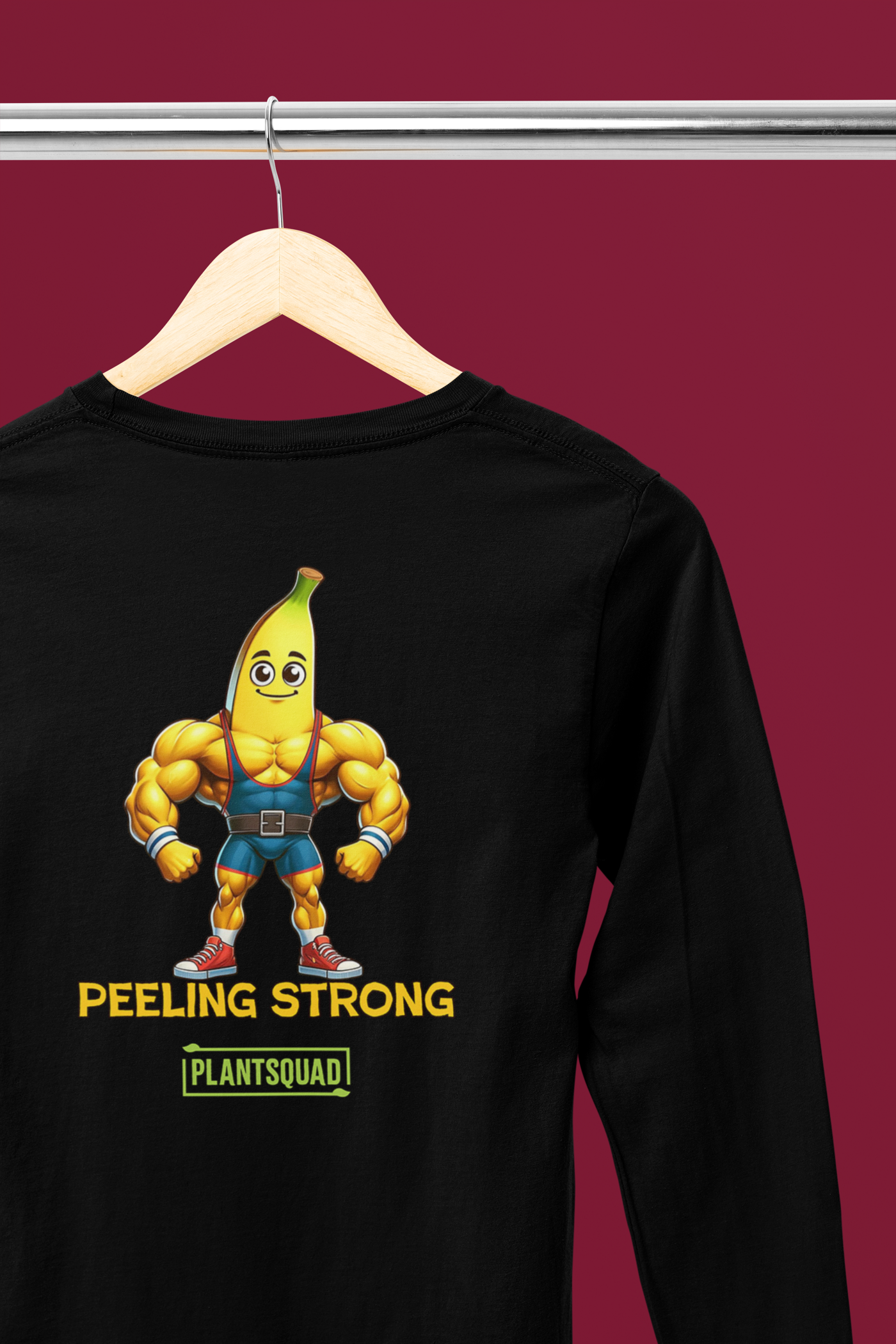 A Plantsquad Banana "Peeling Strong" - Unisex Long Sleeve T-Shirt featuring a muscly banana cartoon with a smiling face and flexing arms. Below the banana, the text reads "PEELING STRONG" and "PLANTSQUAD," perfect for fitness enthusiasts embracing a vegan lifestyle.