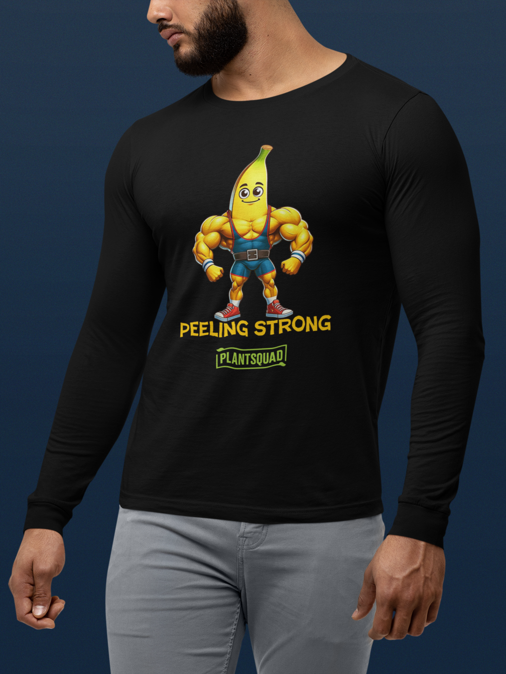 A Plantsquad Banana "Peeling Strong" - Unisex Long Sleeve T-Shirt featuring a muscly banana cartoon with a smiling face and flexing arms. Below the banana, the text reads "PEELING STRONG" and "PLANTSQUAD," perfect for fitness enthusiasts embracing a vegan lifestyle.