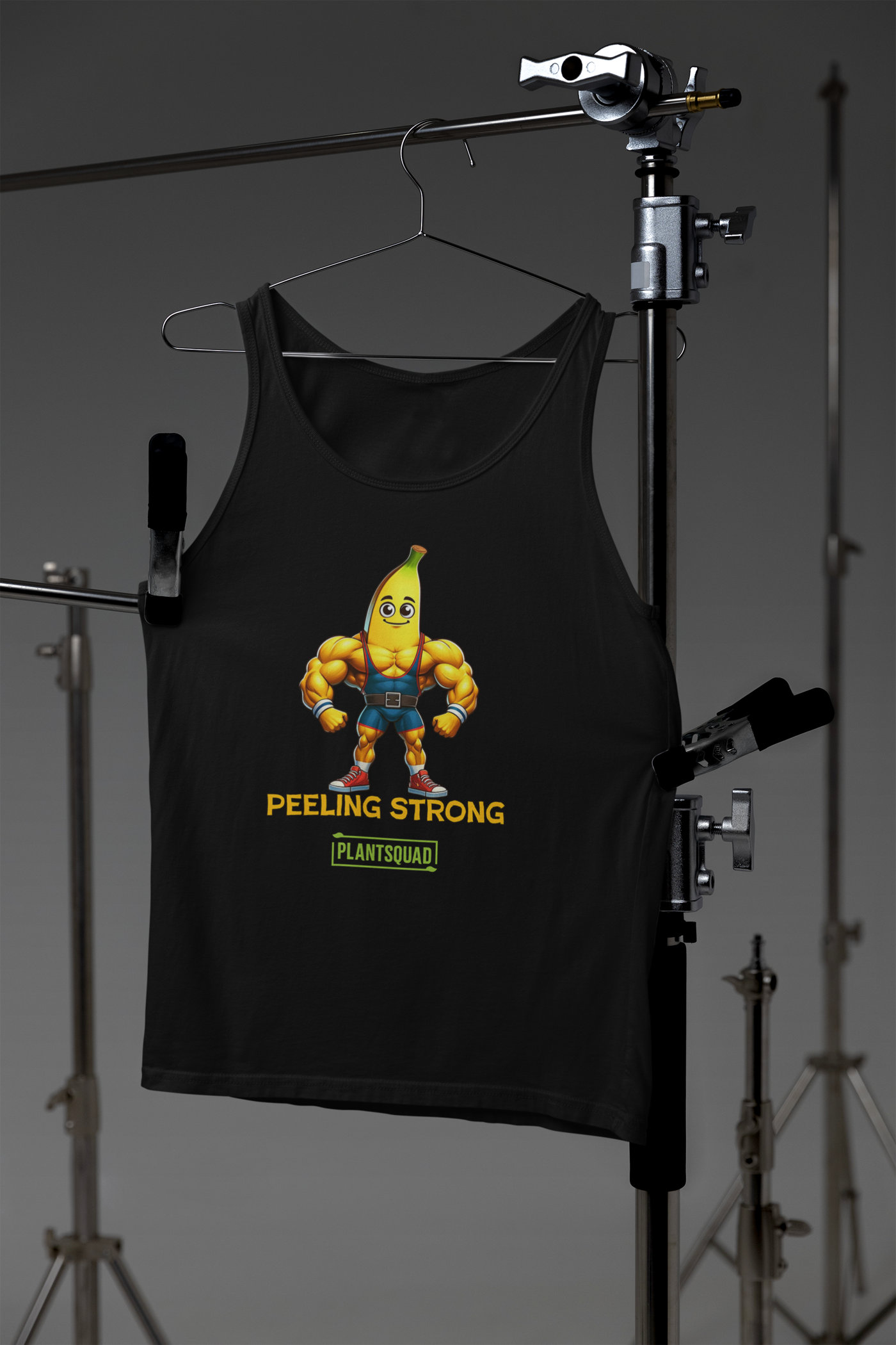 A Plantsquad Banana "Peeling Strong" - Unisex Tank Top features a muscly banana cartoon with a muscular human body, wearing gym clothes and flexing its arms. The text "PEELING STRONG" is written below the image, with "PLANTSQUAD" displayed underneath in a green rectangle, perfect for those embracing a plant-based lifestyle and vegan gym wear.