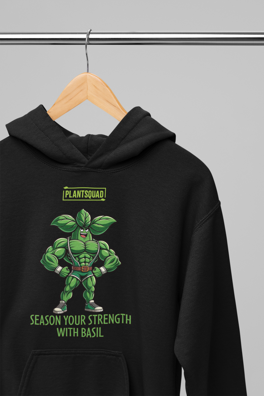 The Plantsquad Basil "Season Your Strength With Basil" - Unisex Hoodie features a muscular cartoon basil character, aptly named Basil, standing with arms crossed. Above Basil, a green logo reads "PLANTSQUAD." Below him, the text says, "SEASON YOUR STRENGTH WITH BASIL." Embrace the vegan lifestyle and cozy fabric while flaunting your love for weight lifting.