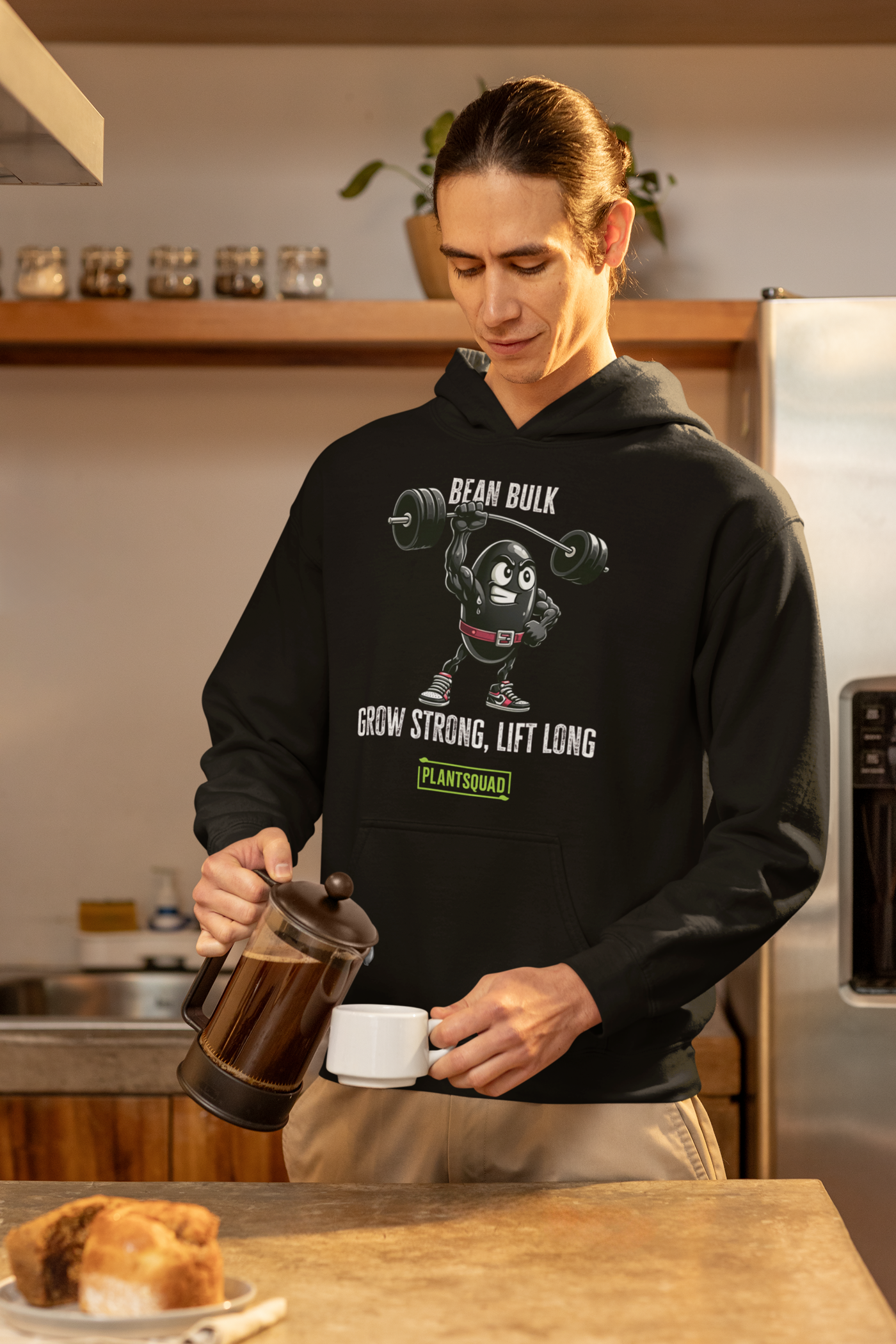 A Plantsquad Black Bean "Bean Bulk Grow Strong Lift Long" - Unisex Hoodie featuring a cartoon bean lifting a barbell with the text "BEAN BULK" above it. Below the bean, the text reads "GROW STRONG, LIFT LONG" and "PLANTSQUAD" within a green rectangle. Perfect for those embracing a vegan lifestyle, this hoodie has a front pocket and drawstrings at the neck.