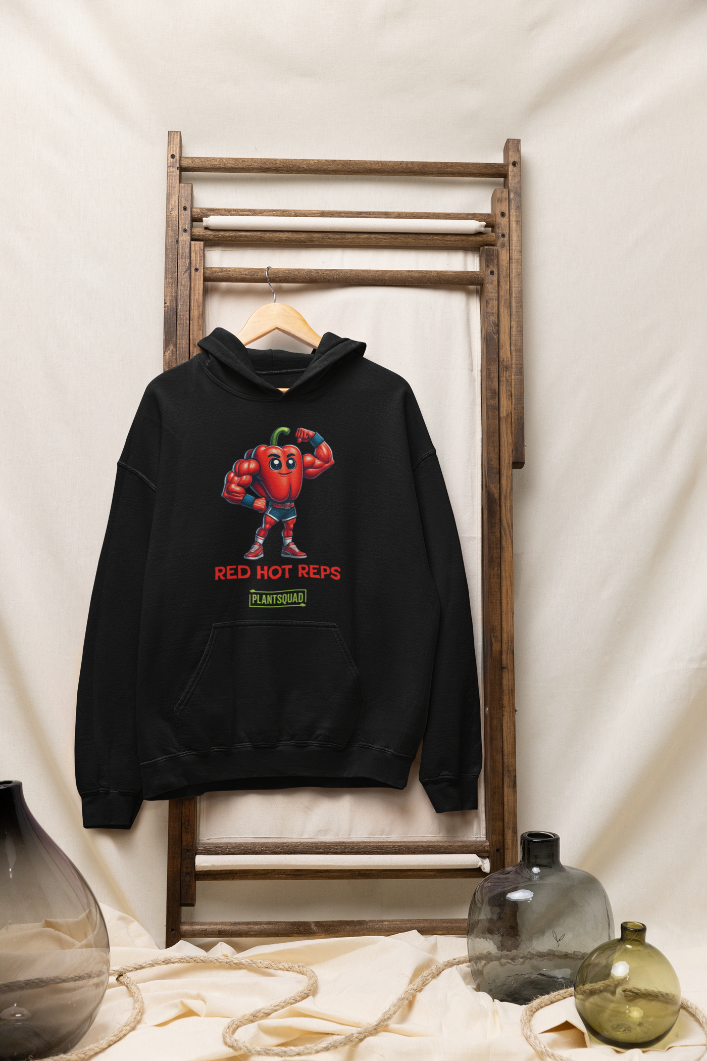 The Plantsquad Capsicum "Red Hot Reps" - Unisex Hoodie features a cartoon muscular red bell pepper character flexing its arms. The text "RED HOT REPS" is printed in red below the character, with "PLANTSQUAD" in green beneath it. Perfect workout clothing, the hoodie has a front pocket and drawstrings at the neck.