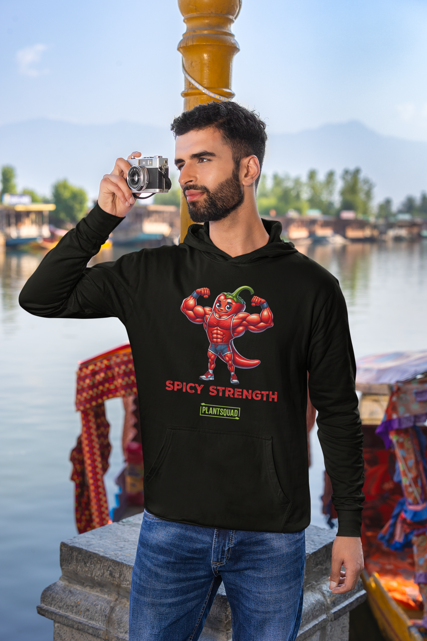 The Plantsquad Chilli "Spicy Strength" - Unisex Hoodie features a cartoon muscular chili pepper character flexing its biceps. The text "SPICY STRENGTH" is written below in red, bold letters. Lower on the hoodie, there is a green rectangular label with the text "PLANTSQUAD" in white, perfect for anyone embracing the vegan lifestyle.