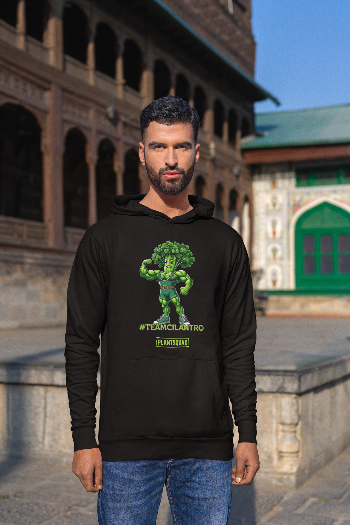 The Plantsquad Cilantro "Team Cilantro" - Unisex Hoodie features a cartoon of a muscular, flexing cilantro superhero, perfect for those embracing a vegan lifestyle. The character has a leafy head, an eye mask, and a belt. Below the character, the text reads "#TEAMCILANTRO" and "PLANTSQUAD.