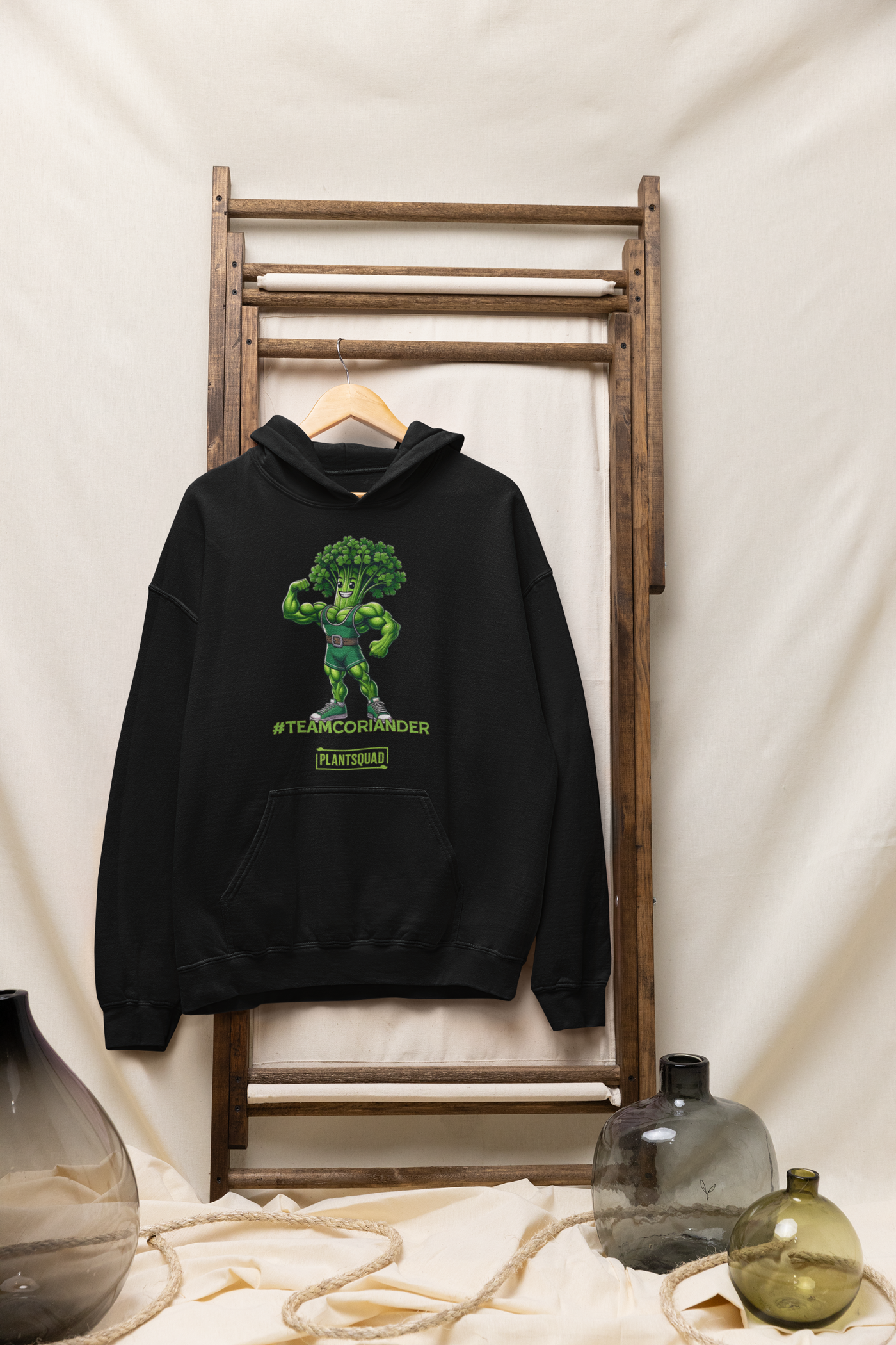 The Plantsquad Coriander "Team Coriander" - Unisex Hoodie features a cartoon illustration of a muscular green stalk of coriander wearing a superhero outfit, flexing its right arm. Embracing the vegan lifestyle, below it are the words "#TEAMCORIANDER" and "PLANTSQUAD" in bold, green text.