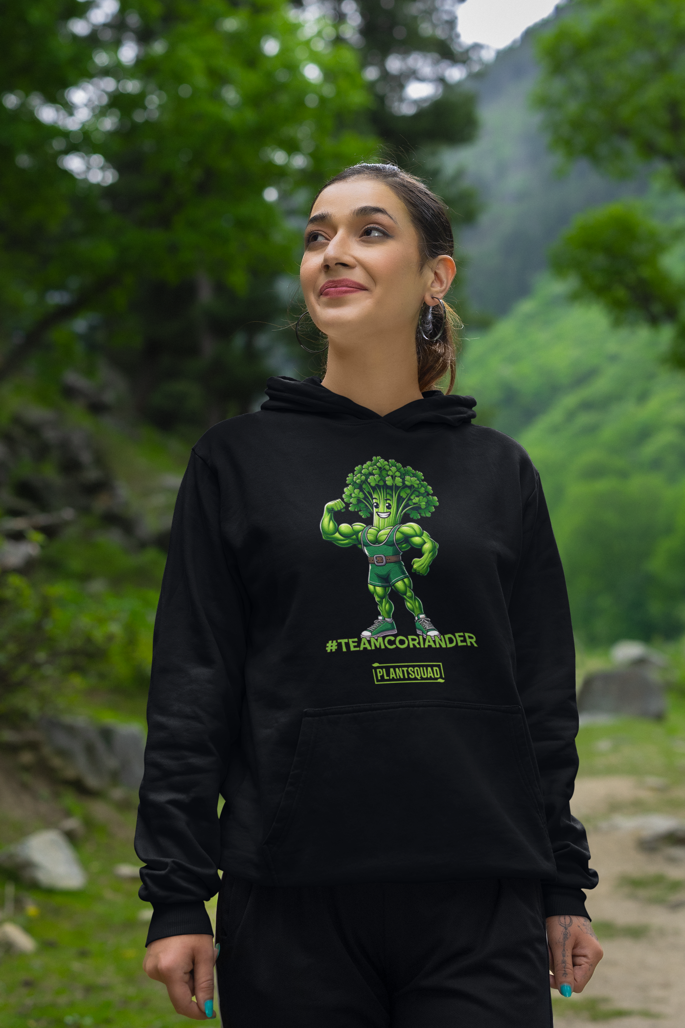 The Plantsquad Coriander "Team Coriander" - Unisex Hoodie features a cartoon illustration of a muscular green stalk of coriander wearing a superhero outfit, flexing its right arm. Embracing the vegan lifestyle, below it are the words "#TEAMCORIANDER" and "PLANTSQUAD" in bold, green text.