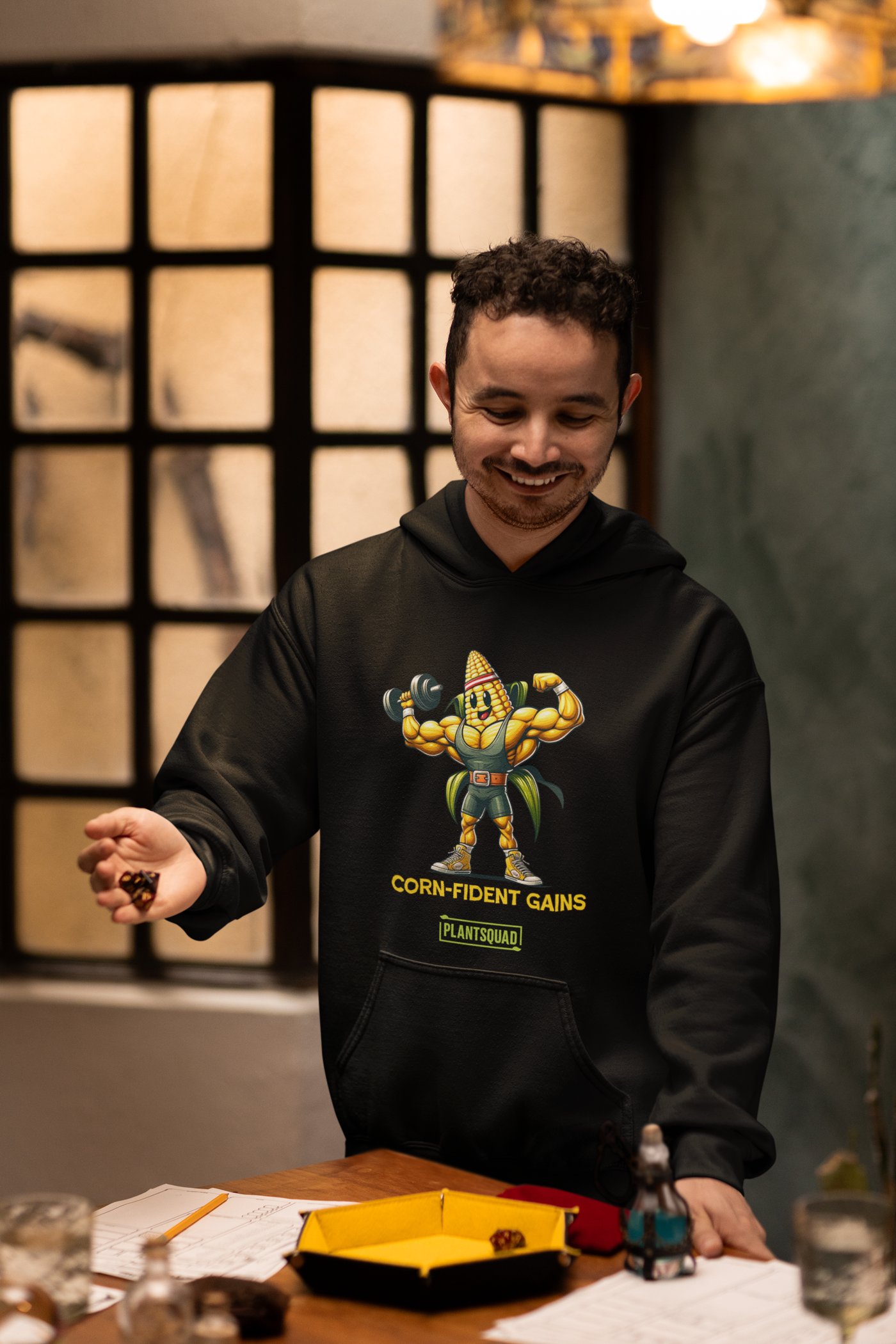 Black hoodie featuring a muscular anthropomorphic corn cob character flexing its biceps. The text beneath the character reads "CORN-fident Gains" with "PLANTSQUAD" in a green box below. Perfect for gym enthusiasts who embrace a plant-based lifestyle, this Plantsquad Corn "Corn-fident Gains" - Unisex Hoodie exudes confidence with every wear.