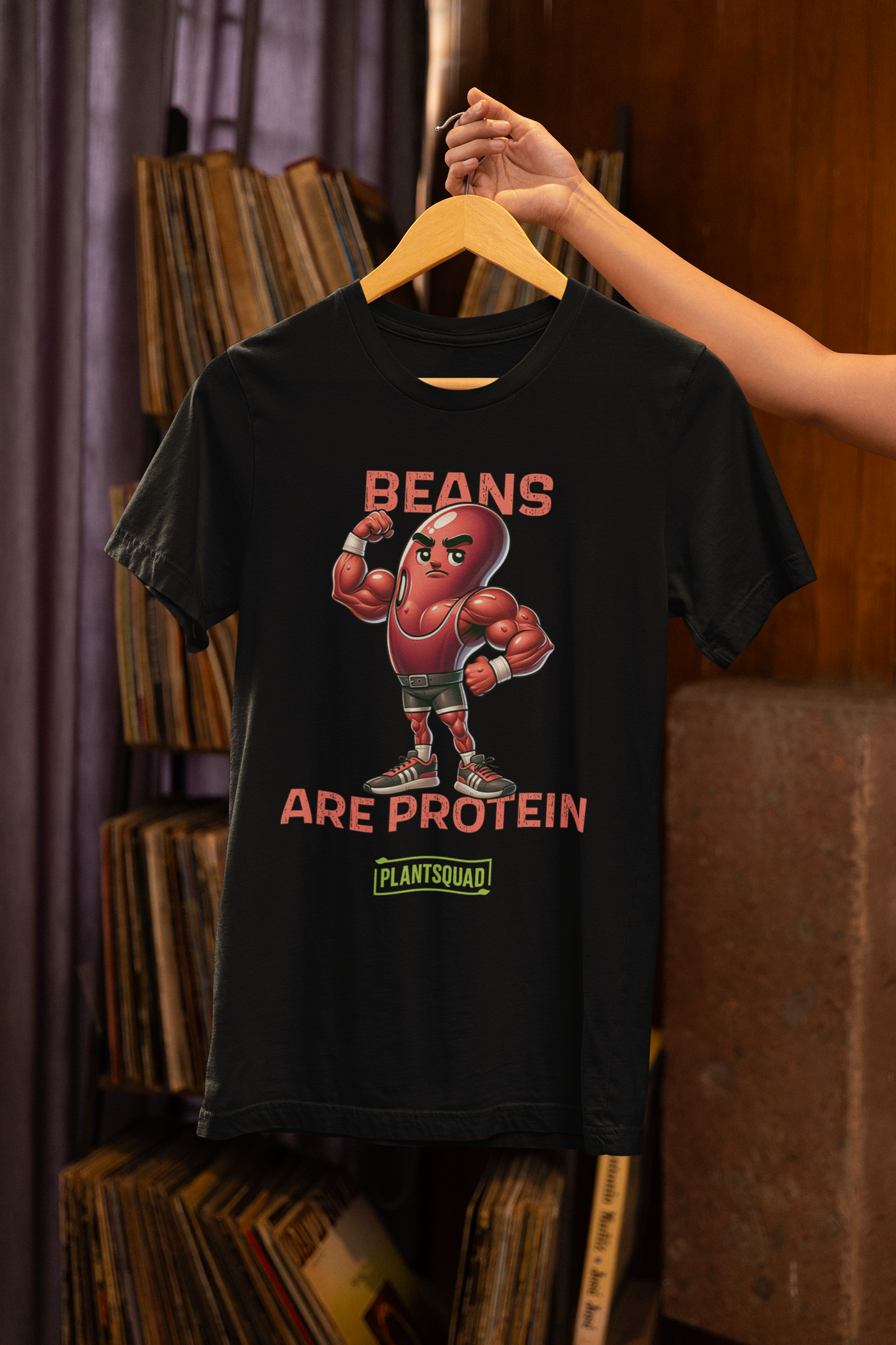 A black t-shirt featuring a muscular cartoon bean character with arms flexed. Above the character, red text reads "Beans," and below it, "Are Protein" is displayed in orange. Beneath that, the word "Plantsquad" is prominently written in green — Plantsquad Kidney Bean "Beans Are Protein" - Unisex T-Shirt.