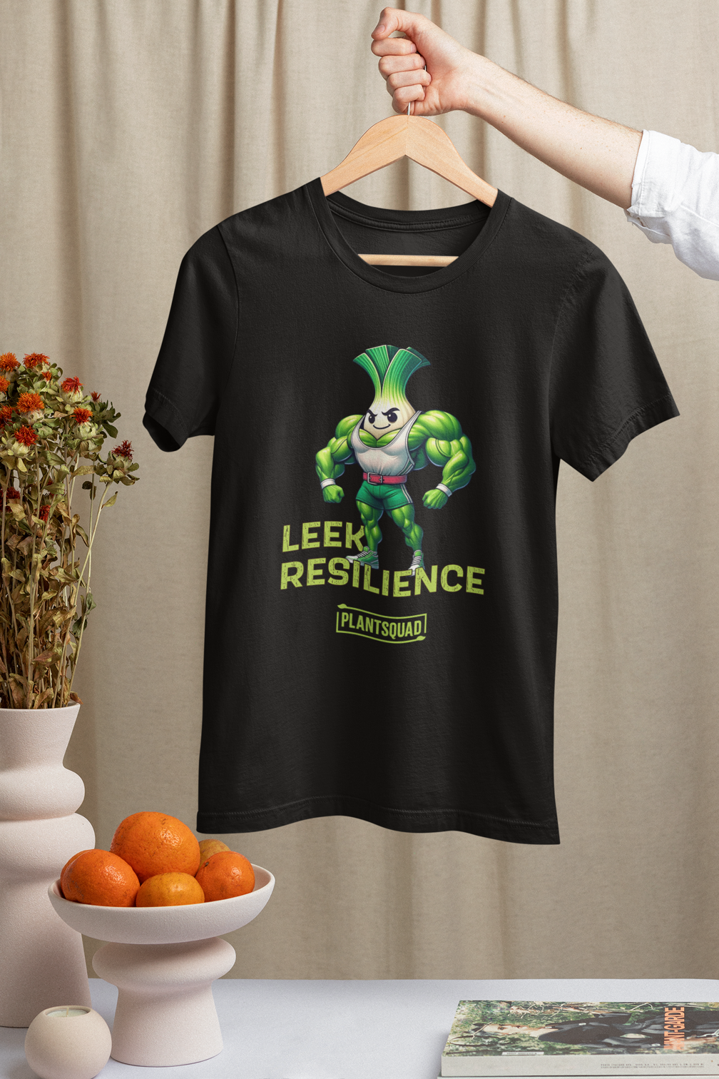 A black T-shirt features a muscular cartoon leek with a determined expression, perfect for embracing a plant-based lifestyle. The leek has a white bulb and green foliage, wearing red shorts. Text reads "Leek Resilience" above and "Plant Squad" below, making it the ultimate fitness t-shirt. Introducing the Plantsquad Leek "Leek Resilience" - Unisex T-Shirt!