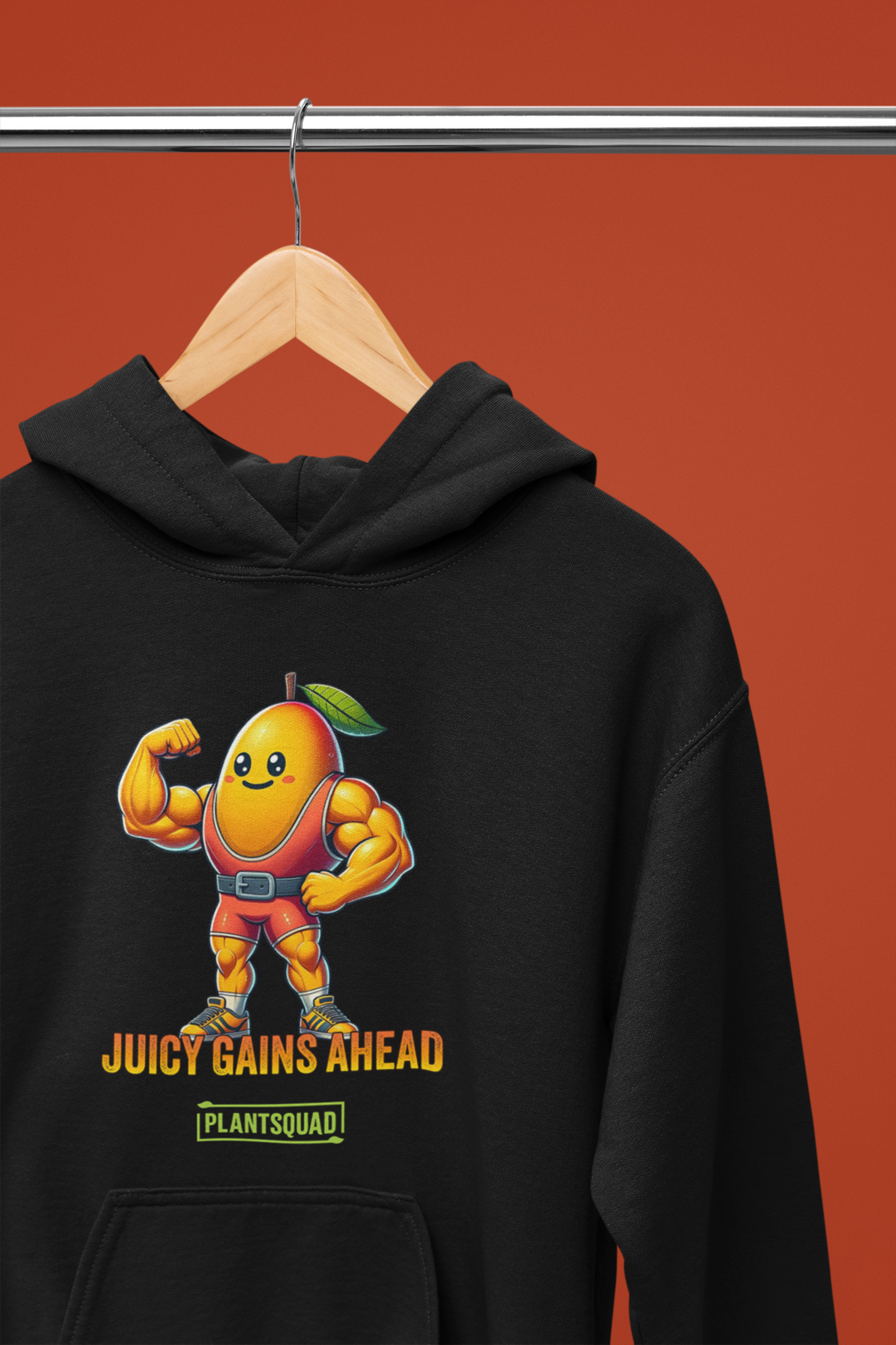 A black Plantsquad Mango "Juicy Gains Ahead" - Unisex Hoodie features a muscular mango cartoon flexing its biceps, donning a green leaf on its head. Below the mango, the text reads "Juicy Gains Ahead" in bold orange letters and "Plant Squad" in a green rectangular box, perfect for showcasing your vegan lifestyle.