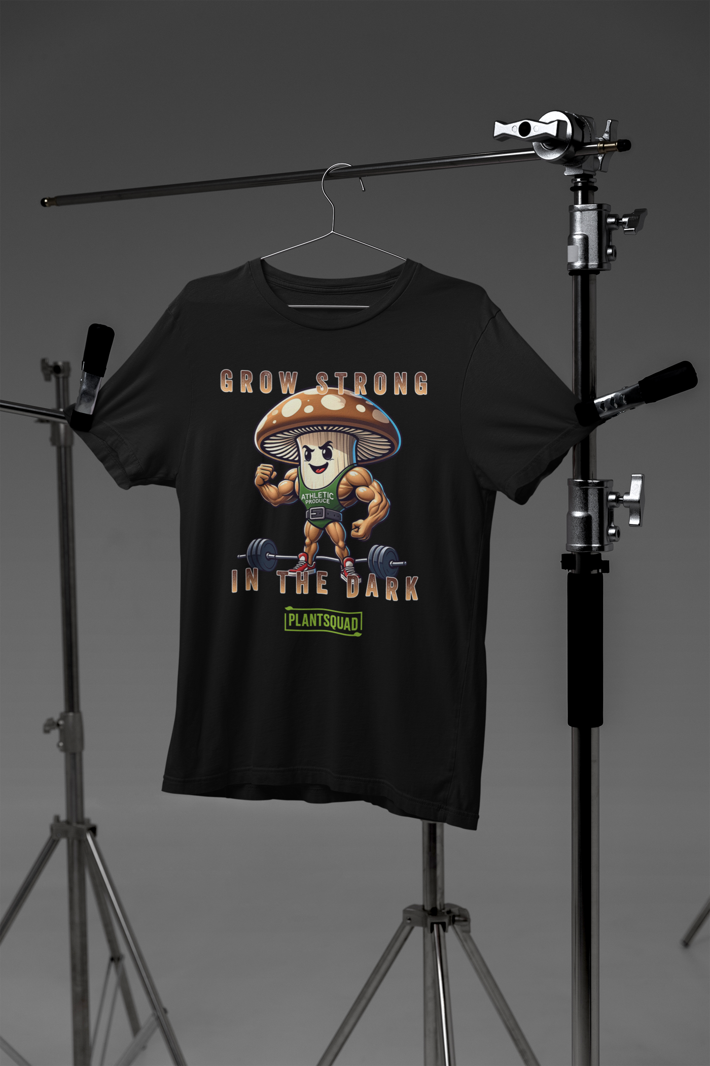 A Plantsquad Mushroom "Grow Strong In The Dark" - Unisex T-Shirt made of breathable fabric featuring a cartoon mushroom character with muscular arms and legs, flexing its biceps and standing in front of a barbell. The text above reads, "GROW STRONG IN THE DARK," and the text below reads, "PLANTSQUAD." Perfect for fitness enthusiasts.