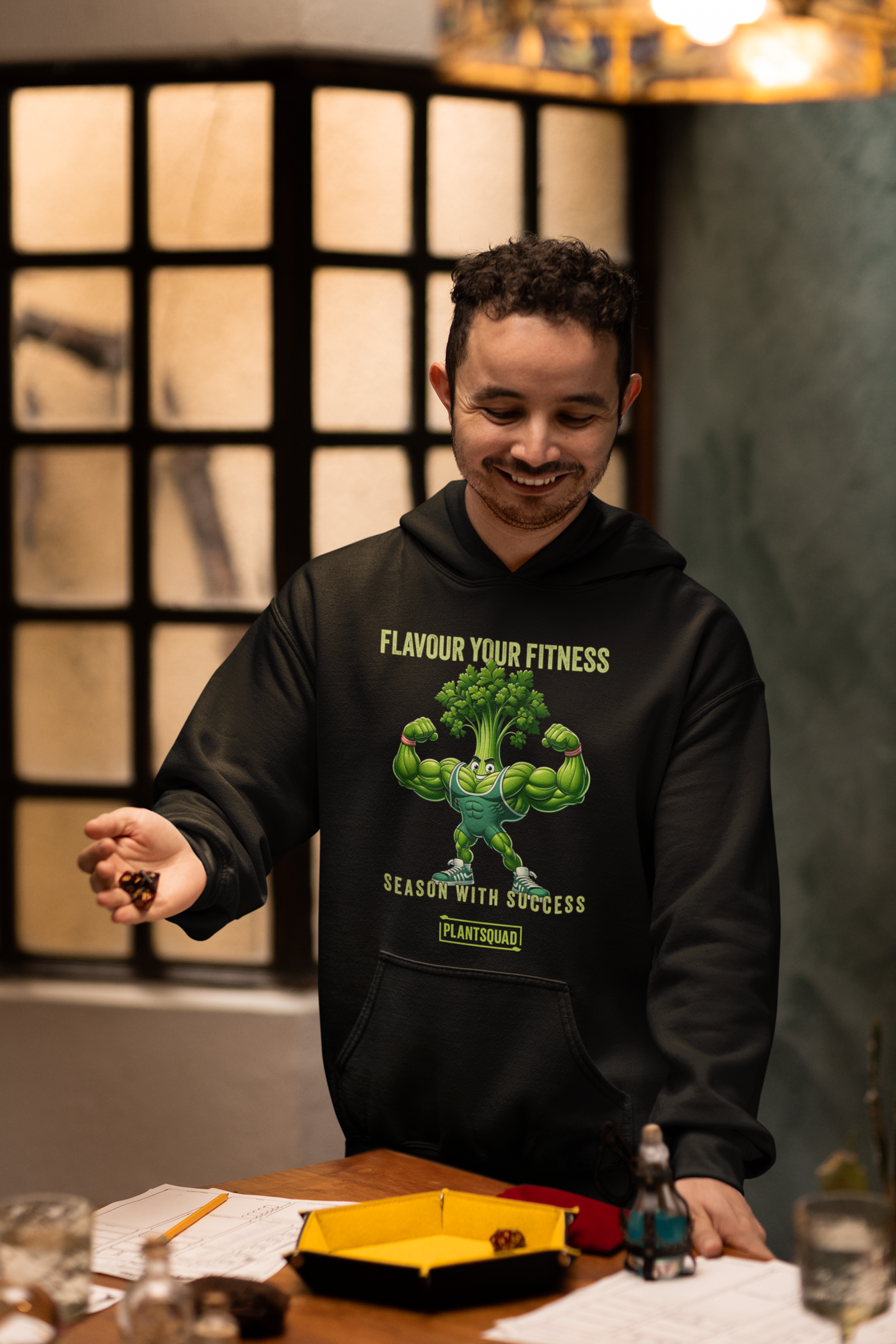 A black vegan hoodie featuring a muscular, anthropomorphic broccoli character flexing its arms. Above the character, the text reads "Flavour Your Fitness," and below it says "Season With Success." The "PLANTSQUAD" logo is displayed at the bottom, perfect for plant-based fitness enthusiasts. This is the Plantsquad Parsley "Flavour Your Fitness Season With Success" - Unisex Hoodie.