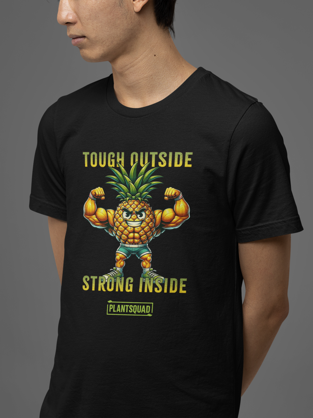 A black workout T-shirt features a muscular anthropomorphic pineapple flexing its biceps. Above the pineapple is the text "Tough Outside" and below it reads "Strong Inside." At the bottom, there's a green box with the word "Plantsquad Pineapple  'Tough Outside Strong Inside' - Unisex T-Shirt," perfect for anyone embracing a plant-based lifestyle.