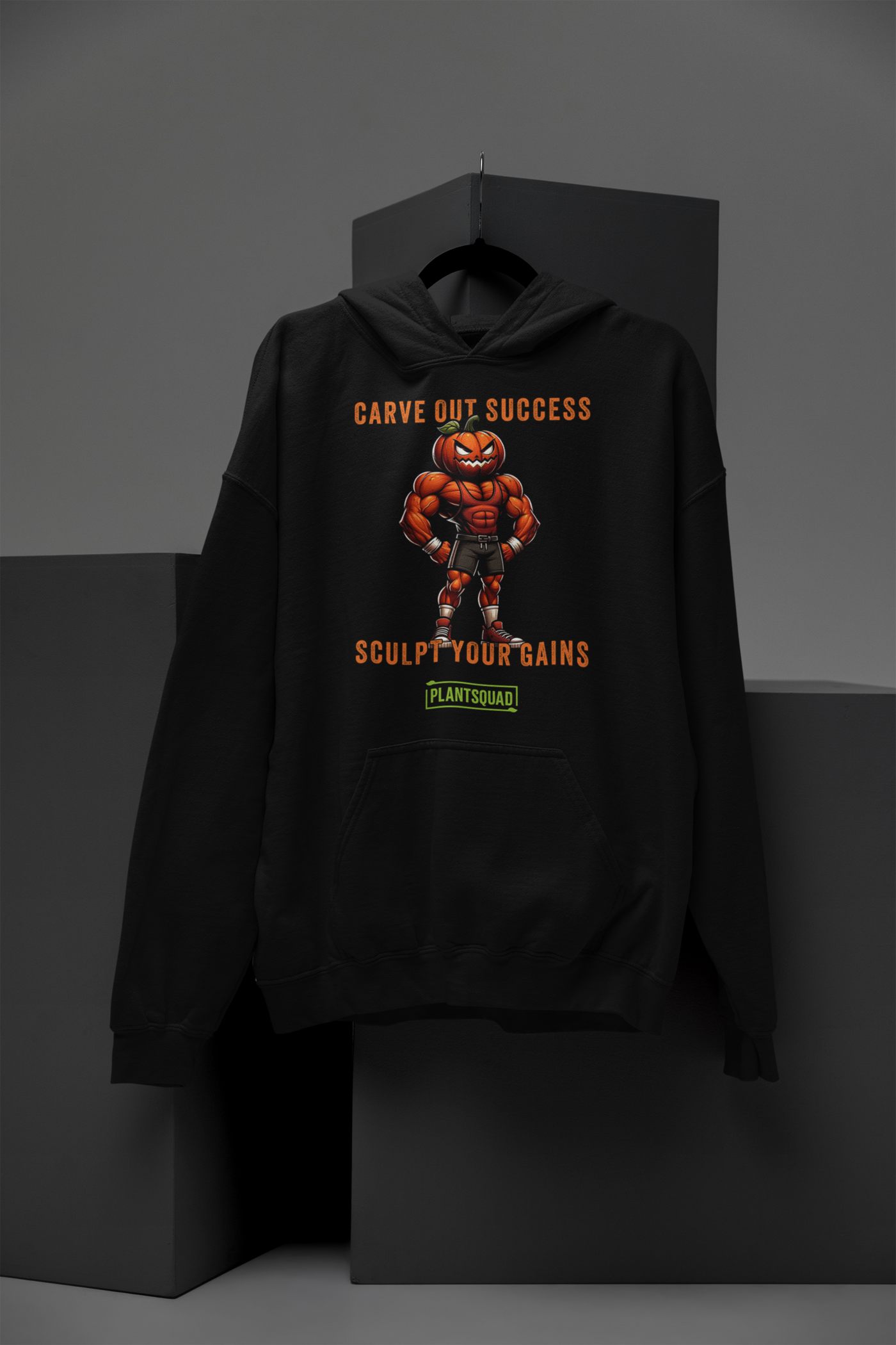 A black weight lifting hoodie features an illustration of a muscular pumpkin-headed figure in workout attire. The text above and below reads "Carve Out Success" and "Sculpt Your Gains," with a small "PlantSquad" logo at the bottom, celebrating the vegan lifestyle. This is the Plantsquad Pumpkin "Carve Out Your Success Sculpt Your Gains" - Unisex Hoodie.