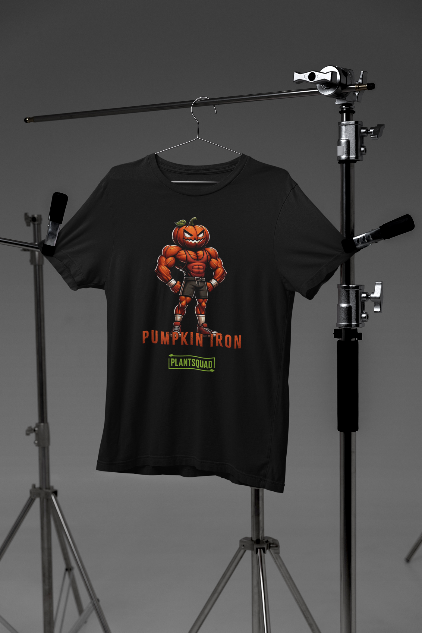 A Plantsquad Pumpkin "Pumpkin Iron" - Unisex T-Shirt featuring a muscular pumpkin-headed character flexing its arms. The character is wearing shorts, trainers, and gloves. The text "PUMPKIN IRON" is displayed in bold orange letters below the character, with "PLANTSQUAD" in green beneath it—perfect for fitness enthusiasts with a vegan lifestyle.