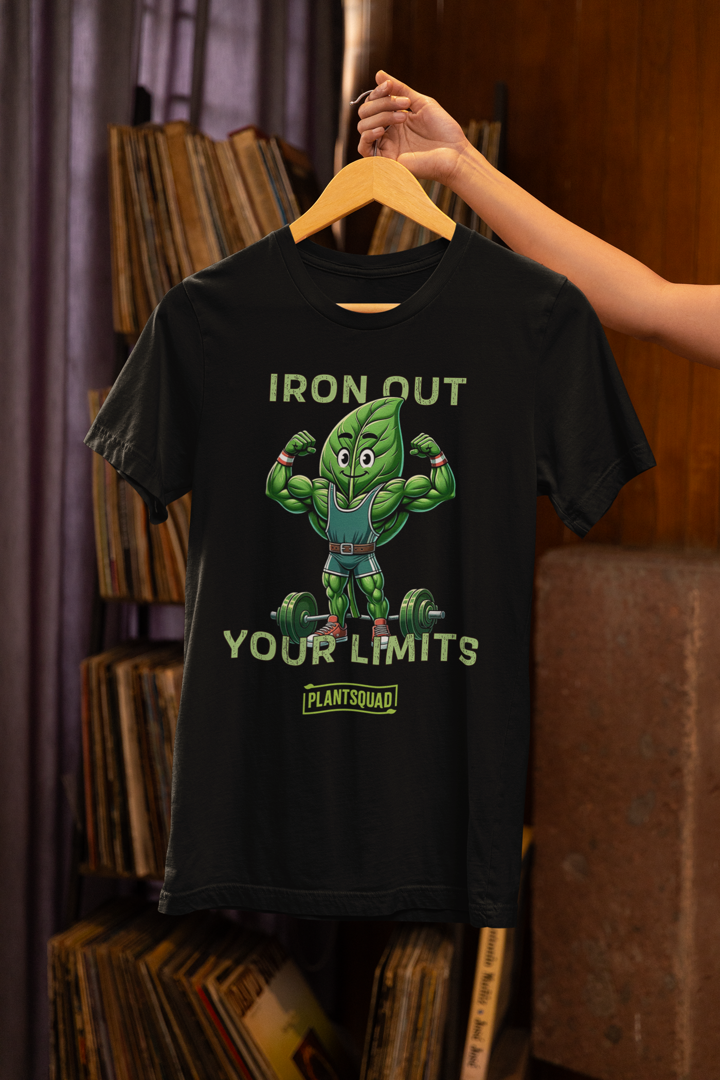 A black Plantsquad Spinach "Iron Out Your Limits" - Unisex T-Shirt featuring a muscular cartoon green leaf character lifting a barbell, with the text "IRON OUT YOUR LIMITS" above and below the character. Embrace your vegan lifestyle with the word "PLANTSQUAD" at the bottom in a small rectangular green box.