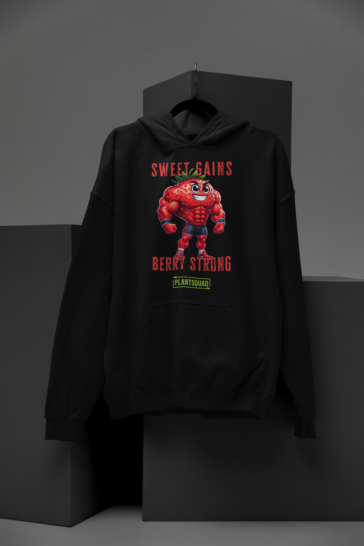 A black Plantsquad Strawberry "Sweet Gains Berry Strong" - Unisex Hoodie featuring a muscular, anthropomorphic strawberry character at the center, perfect for embracing a vegan lifestyle. Text above the strawberry reads "Sweet Gains" and below it reads "Berry Strong" with a small "PlantSquad" logo underneath. Made from cozy fabric, ideal for post-weight lifting comfort.